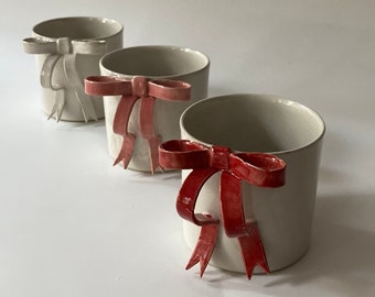 May include: Three white ceramic cups with red ribbon bows. The cups are arranged in a row, with the bow on the leftmost cup facing the viewer, the middle cup's bow facing slightly to the right, and the rightmost cup's bow facing to the right.