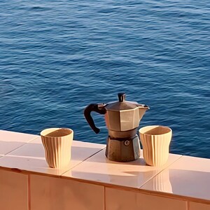 May include: A silver coffee pot with a black handle sits on a pink tiled surface next to two beige ceramic mugs. The background is a blue ocean.