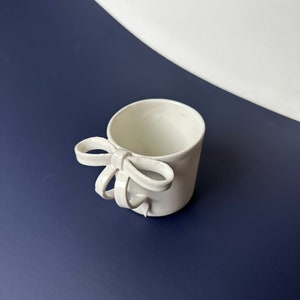 Puede incluir: Taza de cerámica blanca con un asa esculpida en forma de lazo. La taza está sobre una superficie azul.