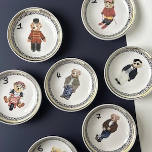 Puede incluir: Juego de siete platos decorativos con ilustraciones de osos con varios atuendos. Cada plato tiene un centro color crema con un borde con estampado negro y dorado. Los osos están representados con atuendos como un esquiador, un motorista y un uniforme militar.