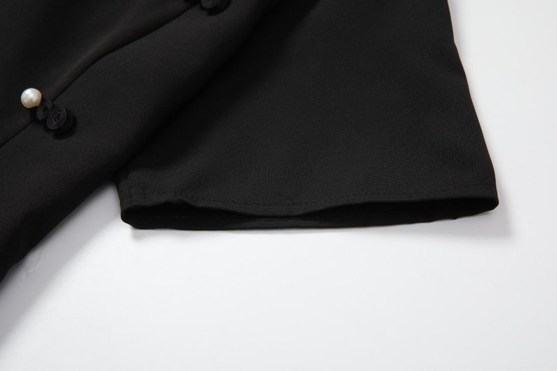 May include: Close-up of the black fabric sleeve of a dress with a pearl button detail.
