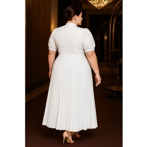 May include: A white midi dress with a high neckline and short, puffed sleeves. The skirt is pleated, and the dress is worn with gold heels. The dress is a solid color and appears to be made of a lightweight fabric.