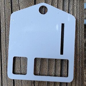 May include: A light gray metal sign with a house-shaped top, a hole at the top, a vertical slit, and two rectangular cutouts. The sign is set against a weathered wooden background.