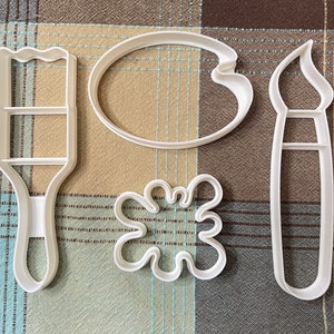 May include: Set of four white plastic cookie cutters shaped like a paintbrush, paint palette, paint splatter, and a paint tube.