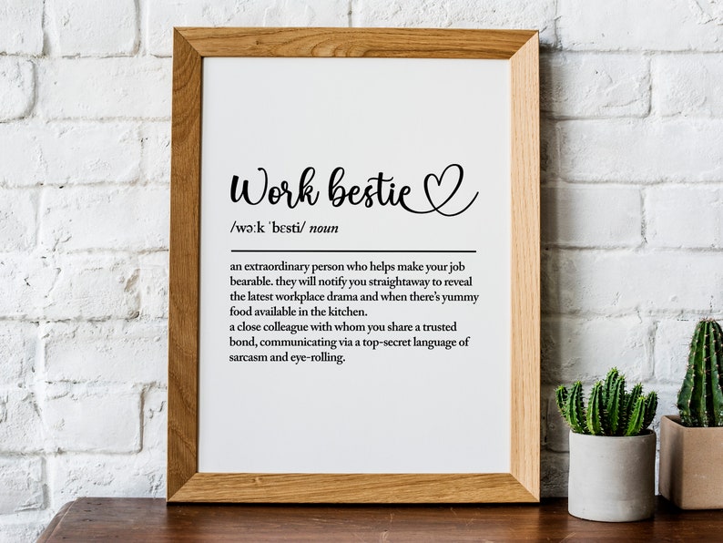 May include: A framed print with a white background and black text that defines "work bestie" as an "extraordinary person who helps make your job bearable." The print is on a wooden table with a small potted cactus in the corner.
