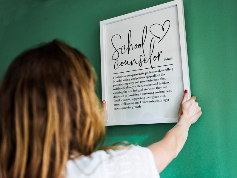 May include: A white framed print with the text "School Counselor" in black cursive lettering. The text is defined as "a skilled and compassionate professional, excelling in multitasking, and possessing qualities like patience, empathy, and resourcefulness. They collaborate closely with educators and families, ensuring the well-being of students. They are dedicated to providing a nurturing environment for all students, supporting their goals with attentive listening and kind words, ensuring a secure space for growth."