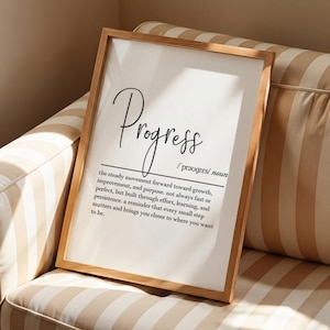 Puede incluir: Un marco de madera clara que contiene una impresión con la palabra "Progress" escrita en cursiva. La definición de abajo describe el movimiento constante hacia el crecimiento, la mejora y el propósito. La impresión está sobre un sofá a rayas beige y blanco.