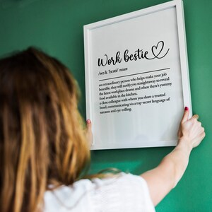 May include: A white framed print with a black and white definition of "work bestie" with a heart symbol. The print is hanging on a green wall.