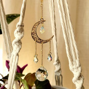 Peut inclure: Un charme en forme de croissant de lune doré avec des accents de cristal suspendu à un porte-plante en macramé. Le charme présente un design délicat en filigrane et est orné de trois gouttes de cristal et d'une plus grosse boule de cristal.