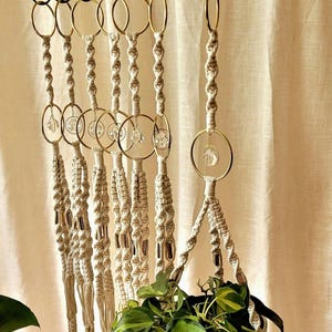 May include: Macrame plant hangers with gold-coloured rings and clear crystal accents. The off-white rope is knotted in a decorative pattern. A green leafy plant is visible in the bottom of the image.