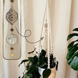 Peut inclure: Un attrape-soleil suspendu composé d'une chaîne en métal doré avec un médaillon en filigrane, une sphère en cristal transparent à facettes suspendue dans un anneau doré, et des breloques en forme de soleil et d'étoile, près d'un porte-plante.