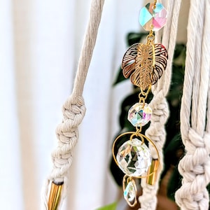 May include: A gold-toned macrame plant hanger with a crystal prism and a gold leaf charm. The hanger is made of natural cotton rope and has a wooden dowel at the top.