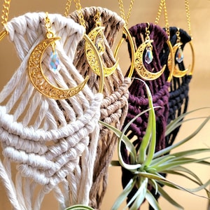 May include: Four macrame plant hangers in cream, tan, dark purple, and black, each featuring a gold-toned metal hoop, a filigree crescent moon charm, and a dangling iridescent crystal bead, suspended by gold-toned chains for display.
