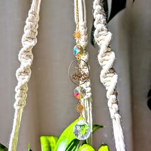 May include: A gold macrame plant hanger with a crystal prism and gold sun charms. The hanger is hanging from a white ceiling and a green plant is in the background.