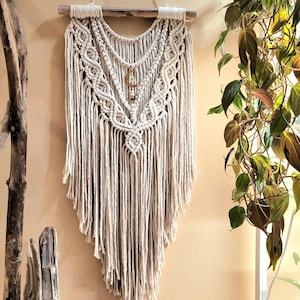 May include: A cream-coloured macrame wall hanging suspended from a horizontal piece of driftwood. The design features intricate knotted patterns, long fringe, and a small gold-toned crescent moon charm centred within the layered cord structure.