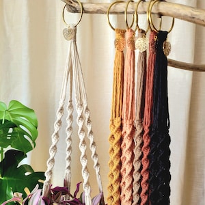 May include: A set of seven macrame plant hangers in various colours, including natural, mustard yellow, peach, light brown, terracotta, and black. Each hanger features a gold leaf charm and is attached to a gold ring.