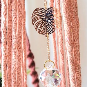 May include: A hanging decoration featuring multiple strands of dusty pink cord. A gold-toned metal monstera leaf charm is suspended from a gold chain, which connects to a clear, multifaceted crystal sphere that reflects light at the bottom.