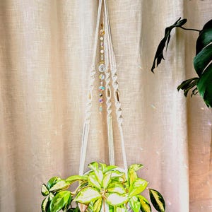 May include: A cream-coloured macrame plant hanger featuring twisted cord patterns and a central vertical strand of multi-coloured glass beads and crystals. The hanger is suspended from a gold-toned metal ring and holds a variegated green and yellow plant.