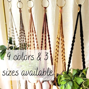 May include: A selection of macrame plant hangers in shades like white, brown, and black. Each hanger, with a metal ring, supports a potted plant. The image includes the text "9 colours & 3 sizes available".