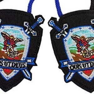 May include: Two black fabric shields with blue trim and embroidered designs. Each shield features a winged angel holding a sword, a banner with the text "Ours Ut Deus", and two crossed swords.