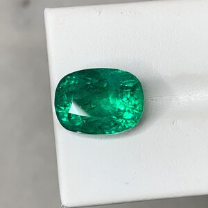 May include: A vibrant green emerald gemstone with a cushion cut. The emerald has a slightly oval shape and is set on a white background.