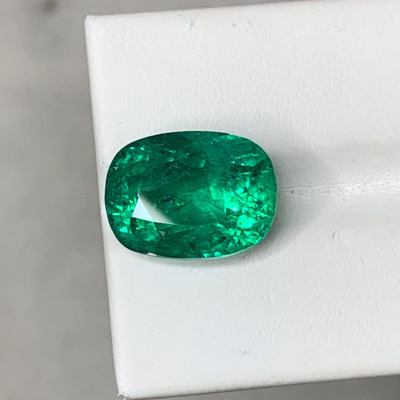 May include: A vibrant green emerald gemstone with a cushion cut. The emerald has a slightly oval shape and is set on a white background.