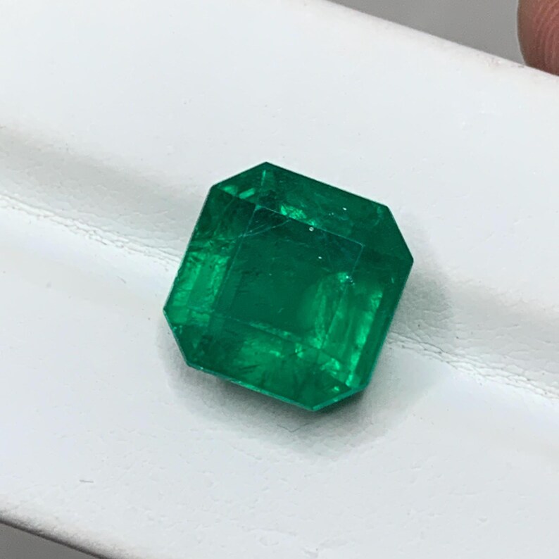 May include: A faceted, emerald-cut gemstone with a vibrant green hue. The gem is displayed on a white surface, showcasing its clarity and brilliance. The emerald's facets reflect light, highlighting its geometric shape and rich color.