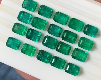 May include: A collection of emerald-cut emerald gemstones. The gemstones are a vibrant green color and are arranged in neat rows. The emeralds are displayed on a white surface, showcasing their clarity and brilliance. The image is well-lit, highlighting the facets of each stone.