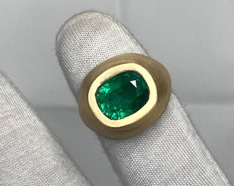 May include: A gold ring featuring a vibrant green emerald gemstone. The emerald is set in a gold bezel, creating a striking contrast. The ring's design is simple yet elegant, showcasing the beauty of the gemstone.