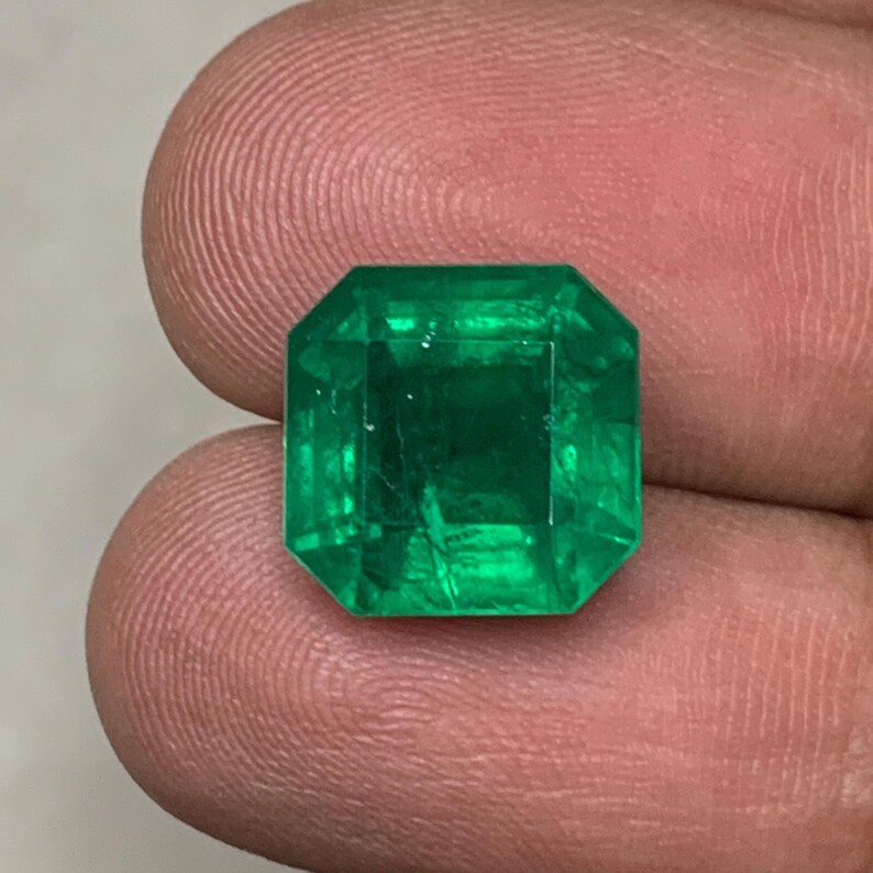 May include: A vibrant, emerald-cut emerald gemstone. The gem is a deep, rich green color with visible internal inclusions. The cut features a rectangular shape with beveled corners, showcasing the stone's clarity and brilliance.
