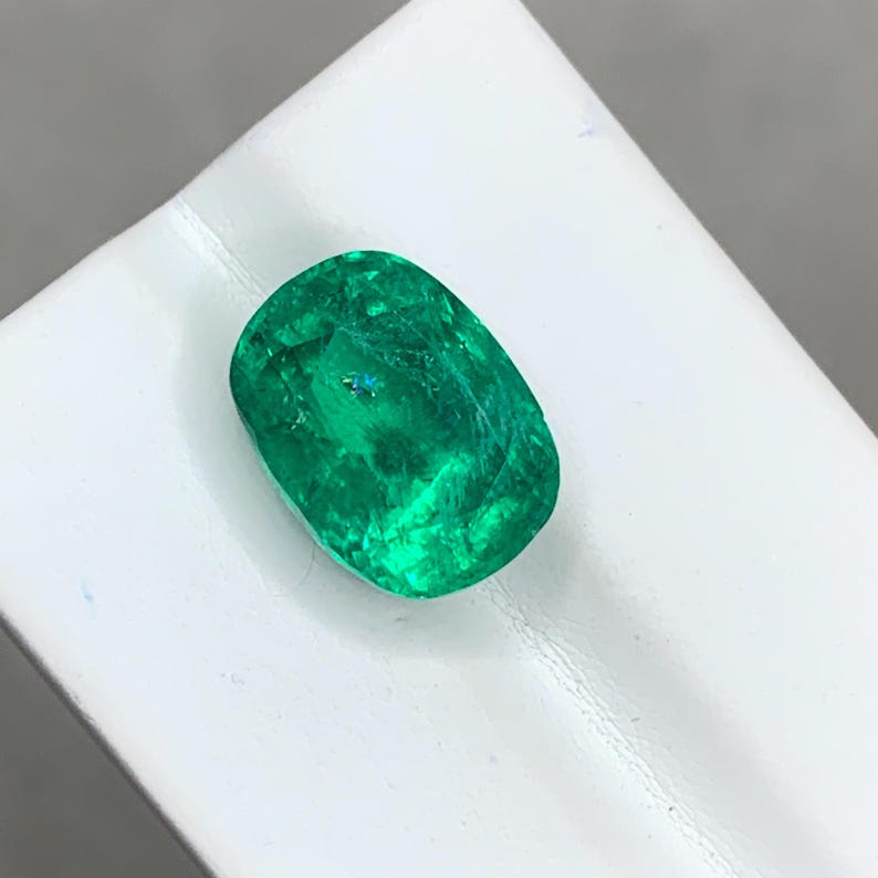 May include: A vibrant green emerald gemstone with a cushion cut. The gemstone is set on a white background.
