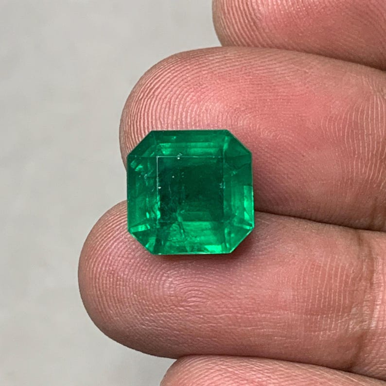 May include: A vibrant, emerald-cut green gemstone. The gem has a square shape with beveled edges, reflecting light. The stone's color is a deep, rich green, and it appears to be faceted to enhance its brilliance. The gemstone is held between fingers.