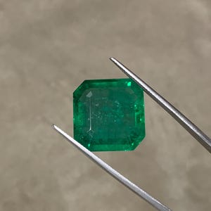 May include: A faceted, emerald-cut emerald gemstone held by tweezers. The gemstone is a vibrant green color and has a rectangular shape with cut corners. The tweezers are silver-colored.