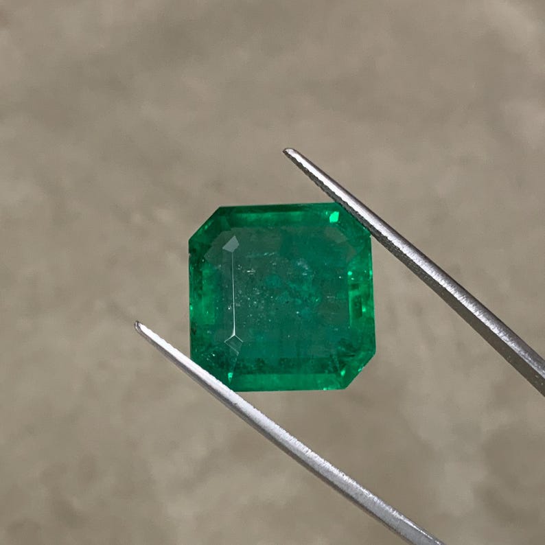 May include: A faceted, emerald-cut emerald gemstone held by tweezers. The gemstone is a vibrant green color and has a rectangular shape with cut corners. The tweezers are silver-colored.