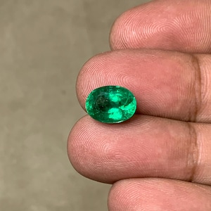 May include: Oval-cut emerald gemstone. The gem is a vibrant green color, with visible inclusions. The stone is held between fingers, showcasing its polished surface and natural characteristics. Ideal for jewelry making or collecting.