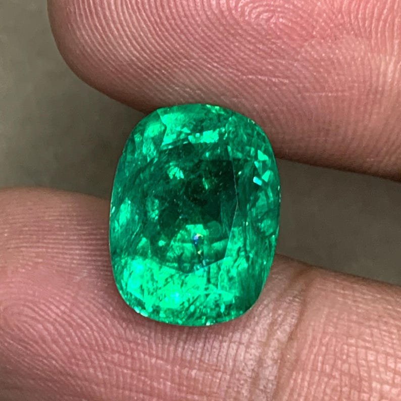 May include: A vibrant green emerald gemstone with a cushion cut. The emerald is set in a ring and is surrounded by a halo of smaller diamonds.