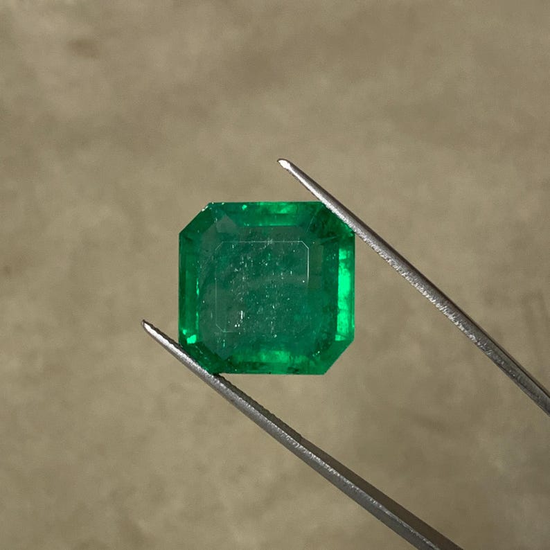 May include: A vibrant, emerald-cut emerald gemstone, held by tweezers. The gem is a deep, rich green color with a square shape and beveled edges. The gemstone is displayed against a neutral background.