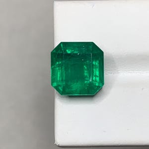 May include: A vibrant, emerald-cut gemstone with a deep green hue. The gem is faceted, showcasing its internal structure and brilliance. The stone is set against a white background, highlighting its color and clarity.