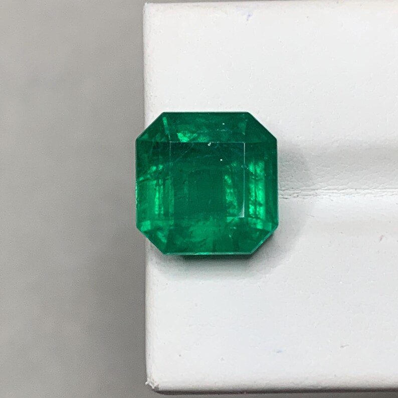 May include: A vibrant, emerald-cut gemstone with a deep green hue. The gem is faceted, showcasing its internal structure and brilliance. The stone is set against a white background, highlighting its color and clarity.