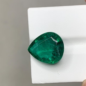 May include: A vibrant, pear-shaped emerald gemstone. The gem is a deep, rich green colour and has a faceted surface that reflects light. The emerald is set against a white background.