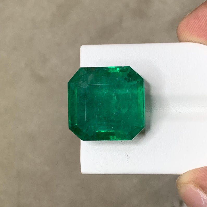 May include: A vibrant, emerald-cut emerald gemstone. The gem is a deep, rich green color and is displayed on a white surface. The emerald has a rectangular shape with cut corners, showcasing its facets.