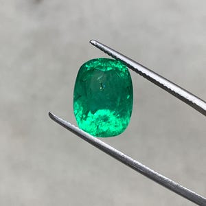May include: A vibrant green emerald gemstone, cut in a cushion shape, is held by a pair of tweezers. The emerald has a deep green color and a slightly cloudy appearance.
