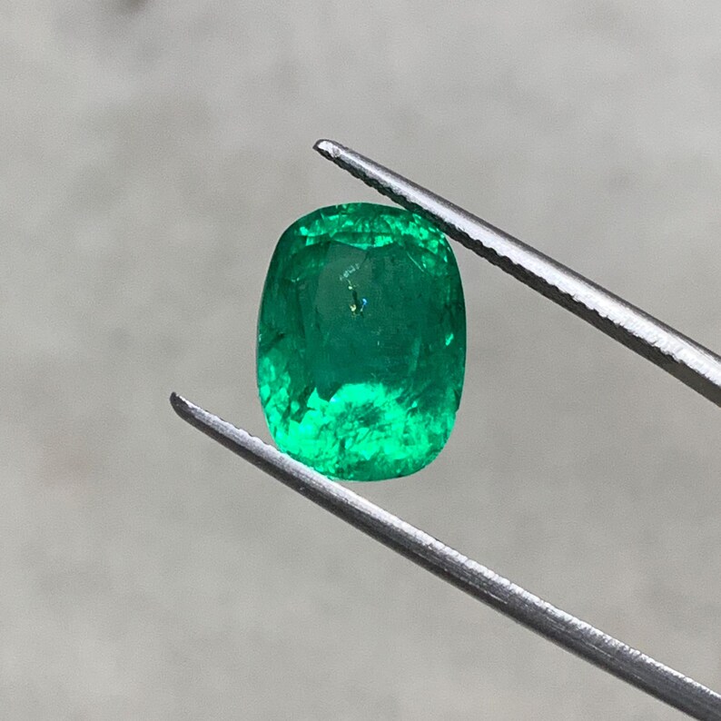 May include: A vibrant green emerald gemstone, cut in a cushion shape, is held by a pair of tweezers. The emerald has a deep green color and a slightly cloudy appearance.