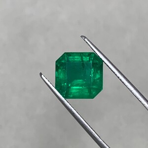 May include: A vibrant, emerald-cut green gemstone held by tweezers. The gemstone has a square shape with beveled edges, showcasing its facets. The gem is a deep, rich green color, reflecting light.