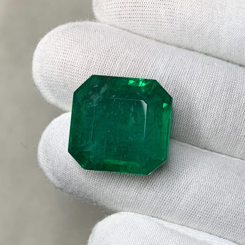 May include: A vibrant, emerald-cut emerald gemstone. The gem is a deep, rich green color with a faceted surface. The emerald is held against a white background.