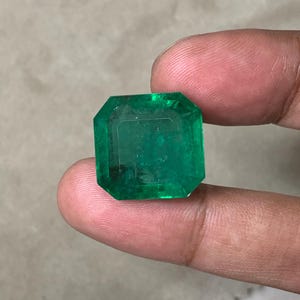 May include: A large, emerald-cut emerald gemstone. The gem is a vibrant green color and has a rectangular shape with beveled edges. The gemstone is held between two fingers.