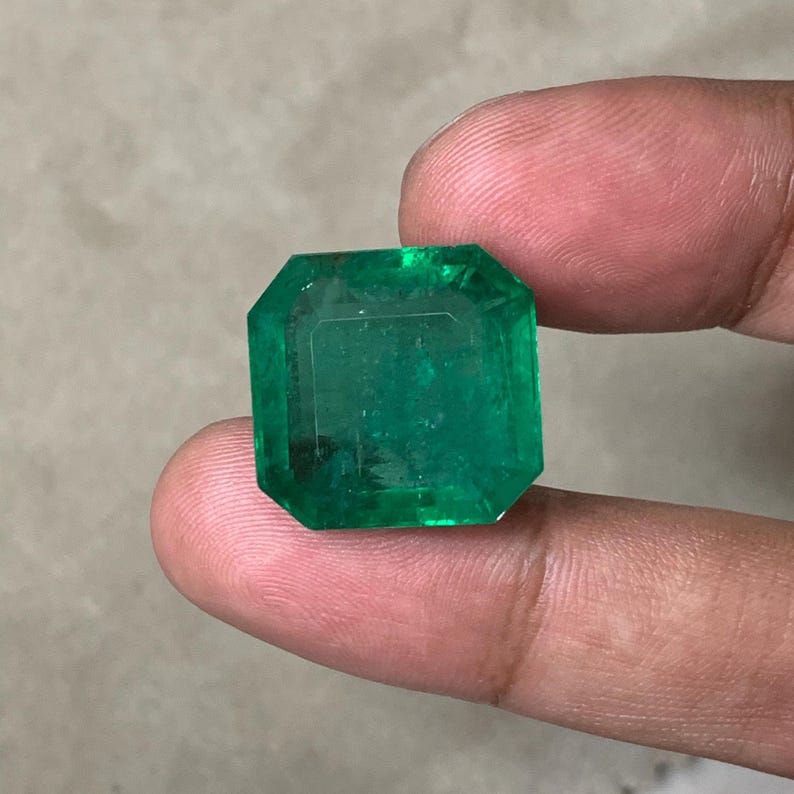May include: A large, emerald-cut emerald gemstone. The gem is a vibrant green color and has a rectangular shape with beveled edges. The gemstone is held between two fingers.