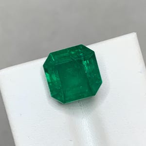 May include: A vibrant, emerald-cut green gemstone. The gem is a deep, saturated green color and appears to be faceted with a rectangular shape. The gemstone is displayed on a white surface.