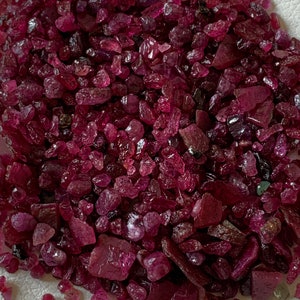 May include: A pile of raw, uncut ruby gemstones. The gems are a deep, rich red color, with varying shapes and sizes. The rough texture of the stones is visible, suggesting their natural state. These gemstones are suitable for jewelry making.