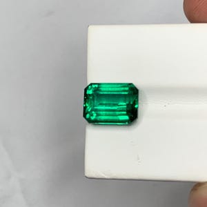 May include: A vibrant, emerald-green gemstone cut in a classic rectangular emerald shape. The stone features sharp, clean facets that reflect light, creating a brilliant, translucent appearance with deep green saturation throughout the entire crystal.